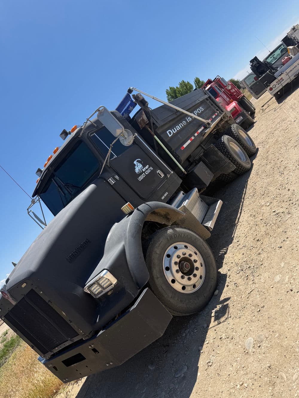 Black dump truck with blue features parked on gravel beside red truck in sunny outdoor setting.