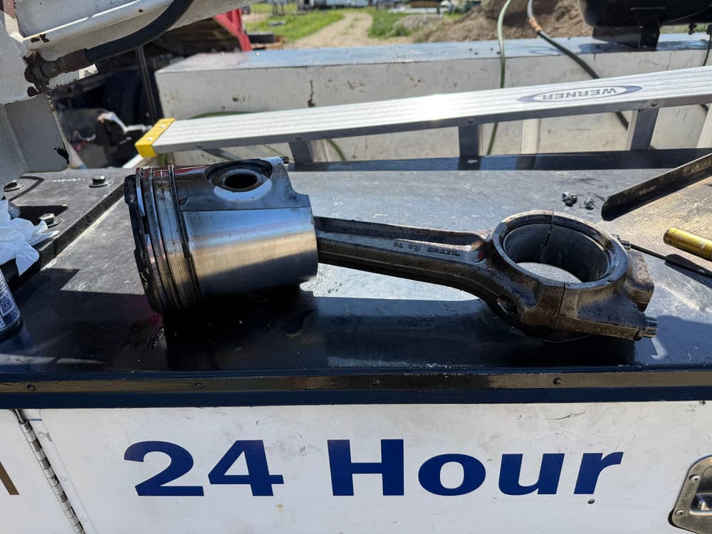 Piston and connecting rod on a workbench with 24 Hour service sign in background.