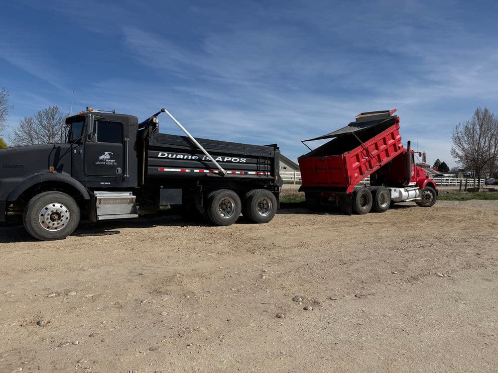 Dump truck unloading gravel on a construction site with clear blue sky and trees in background.