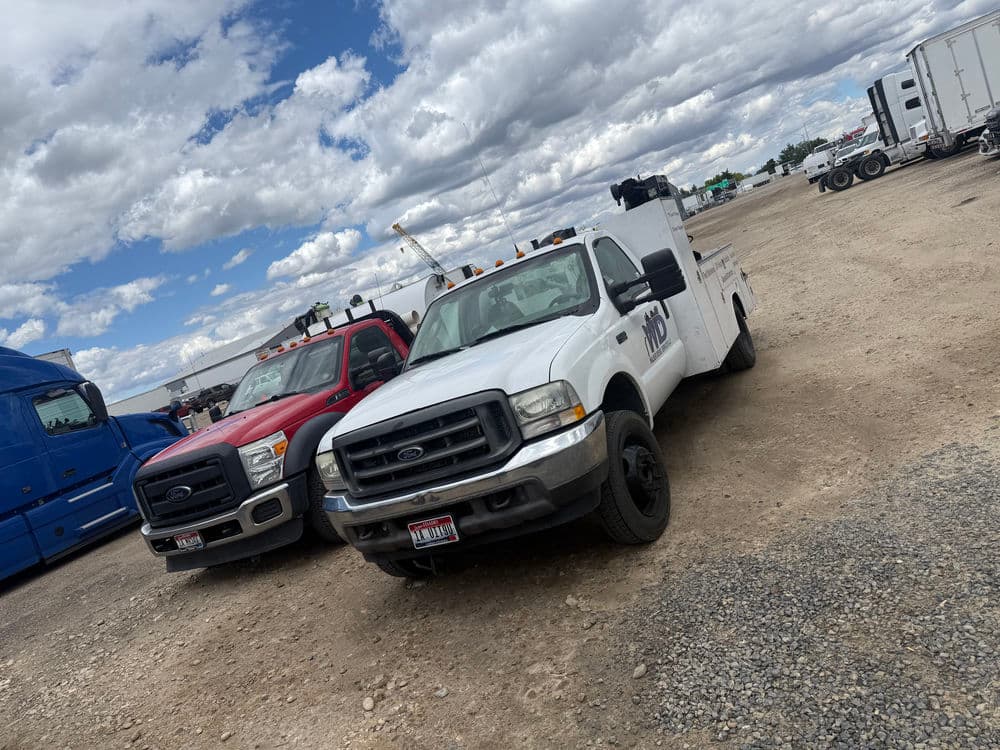 Two work trucks in a gravel lot under a cloudy sky, one white and one red Ford.