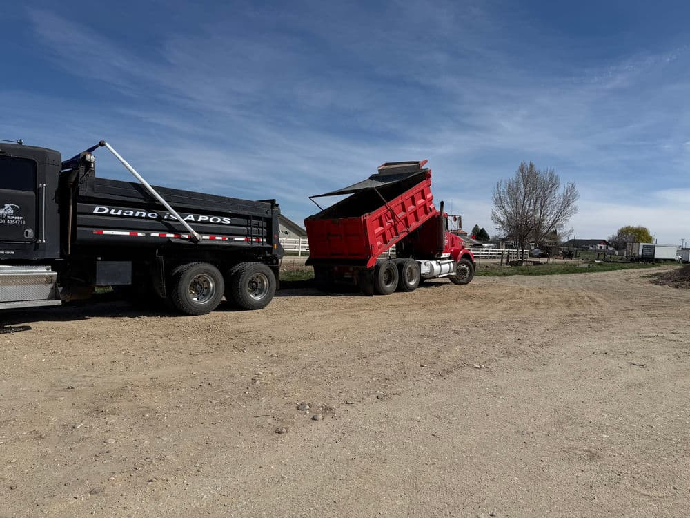 Red dump truck unloading material on a rural road near farm buildings and trees.